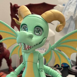 May include: A green and yellow dragon doll with large, round eyes and a toothy grin. The dragon has wings and horns, and is posed standing with its legs slightly bent.