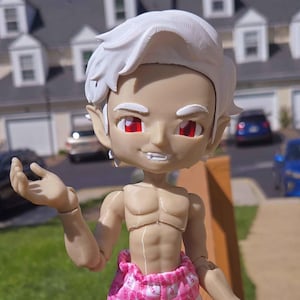 May include: A white-haired doll with red eyes and a toothy grin, wearing pink and white patterned pajamas. The doll has a sculpted, muscular torso and is posed with one arm outstretched.