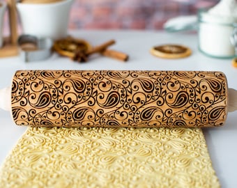 May include: A wooden rolling pin with a paisley pattern engraved into the surface. The rolling pin is being used to roll out dough, which is imprinted with the paisley pattern.