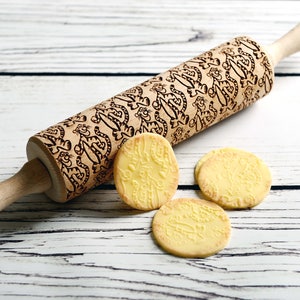 May include: A wooden rolling pin with a carved design of cartoon tigers. Three cookies with the same design are on a white wooden surface.