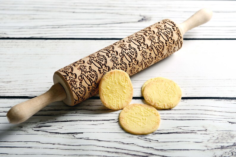 May include: A wooden rolling pin with a carved design of cartoon tigers. Three cookies with the same design are on a white wooden surface.