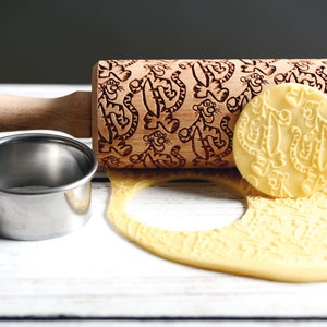 May include: A wooden rolling pin with a carved design of cartoon tigers. The rolling pin is on a white wooden surface with a piece of dough that has been rolled with the pin. There is also a metal cookie cutter on the surface.