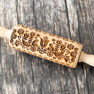 May include: A wooden rolling pin with a carved design of birds and flowers. The design is in a folk art style.