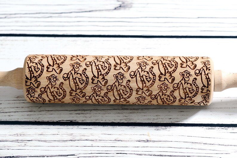May include: A wooden rolling pin with a repeating pattern of cartoon tigers. The tigers are brown and have black stripes. The rolling pin is a light brown color.