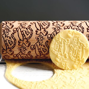 May include: A wooden rolling pin with a carved design of cartoon tigers. The rolling pin is next to a piece of dough with the same tiger design imprinted on it.