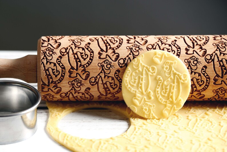 May include: A wooden rolling pin with a carved design of cartoon tigers. The rolling pin is next to a piece of dough with the same tiger design imprinted on it.