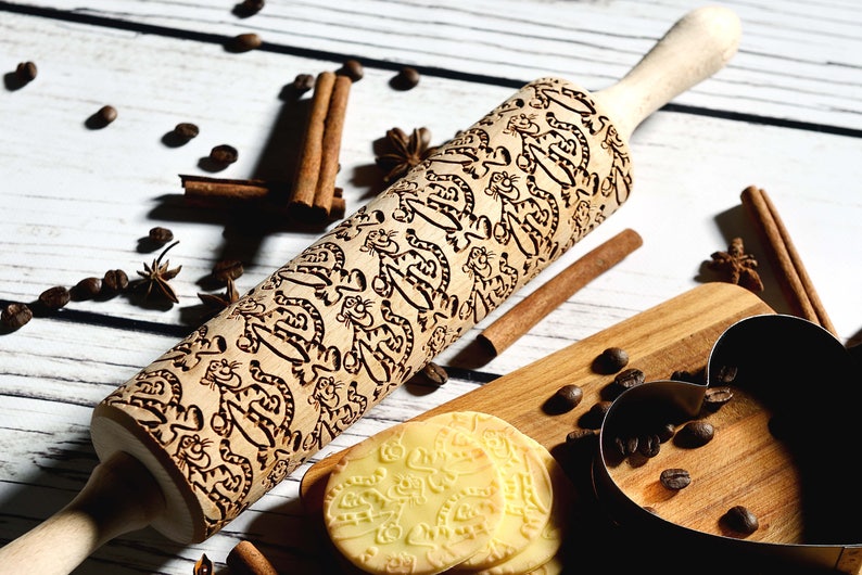 May include: A wooden rolling pin with a carved design of cartoon tigers. The rolling pin is on a white wooden surface with coffee beans, cinnamon sticks, and star anise.
