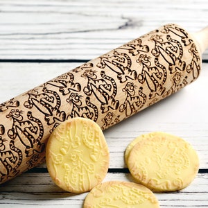 May include: A wooden rolling pin with a carved design of cartoon tigers. The rolling pin is on a white wooden surface with four cookies that have been rolled with the pin.