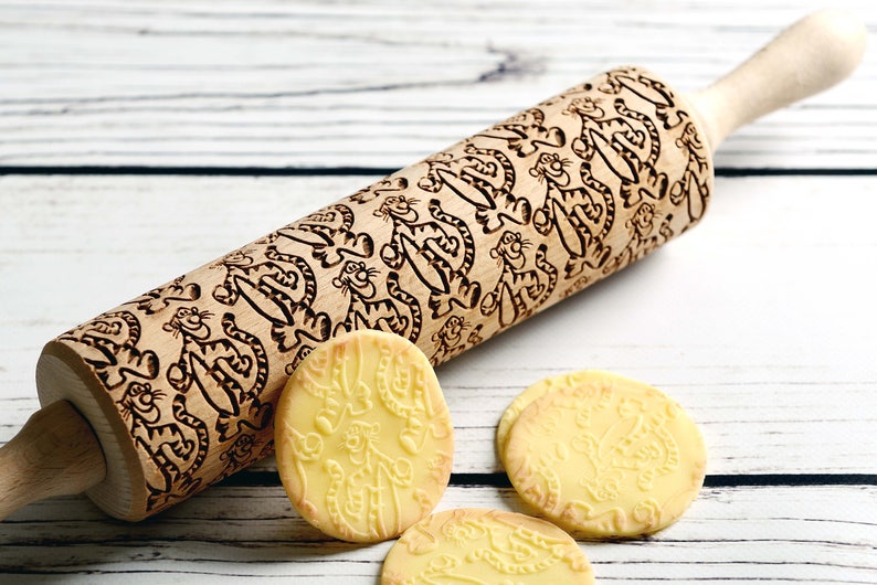 May include: A wooden rolling pin with a carved design of cartoon tigers. The rolling pin is on a white wooden surface with four cookies that have been rolled with the pin.