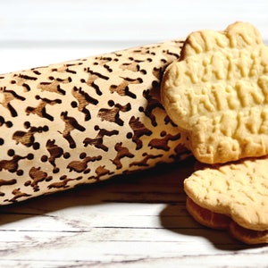 May include: A wooden rolling pin with a carved design of various animals. Two cookies with the same animal design are shown next to the rolling pin.