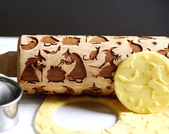 May include: A wooden rolling pin with a Halloween theme. The rolling pin is engraved with images of witches, bats, cauldrons, and stars. A small piece of dough is being rolled out with the rolling pin, showing the design.