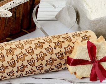 May include: A wooden rolling pin with a repeating pattern of owls engraved on the surface. A cookie with the same owl pattern is in the foreground, tied with a red ribbon.