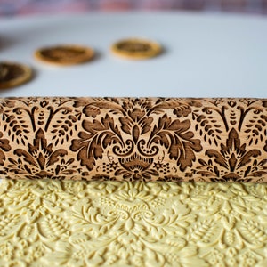 Può includere: Un mattarello di legno con un motivo floreale intagliato. Il mattarello viene utilizzato per creare un design in rilievo su un foglio di pasta gialla.