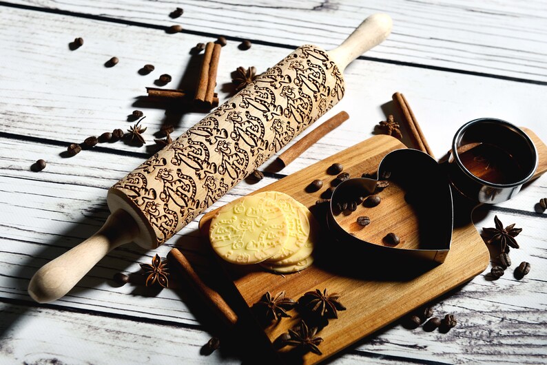 May include: A wooden rolling pin with a decorative engraved pattern, a heart-shaped cookie cutter, and a stack of cookies on a wooden cutting board. The board is surrounded by coffee beans, cinnamon sticks, and star anise.