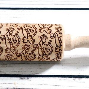 May include: A wooden rolling pin with a brown and black engraved pattern of cartoon tigers. The rolling pin has a wooden handle.