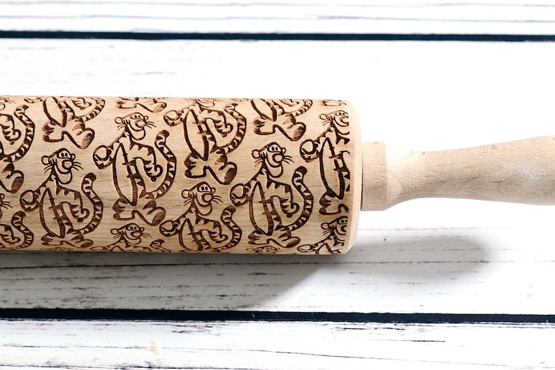 May include: A wooden rolling pin with a brown and black engraved pattern of cartoon tigers. The rolling pin has a wooden handle.