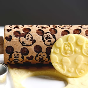 Può includere: Un mattarello di legno con un design di Topolino. Il mattarello è inciso con più immagini della testa di Topolino. Il mattarello è su una superficie bianca con un impasto per biscotti giallo con design di Topolino.