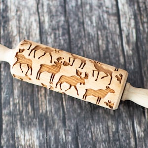 May include: A wooden rolling pin with a carved design of multiple moose and deer. The rolling pin is made of light brown wood and has two handles.