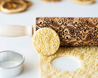 May include: A wooden rolling pin with a decorative paisley pattern engraved into the surface. The rolling pin is shown next to a piece of dough that has been rolled with the pin, and a round cookie cutter.
