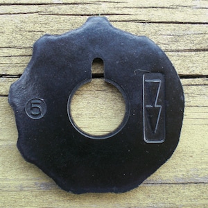 May include: A black, circular sewing machine part with a central hole and a notched outer edge. The number "5" is embossed on the left side, and a symbol is on the right. The part is made of a textured material.
