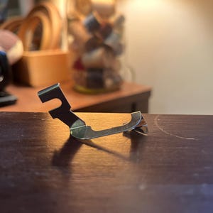 May include: A silver-colored sewing machine presser foot, designed with a curved shape and a notched end. The foot is made of metal and sits on a dark wooden surface, with a blurred background of sewing supplies.