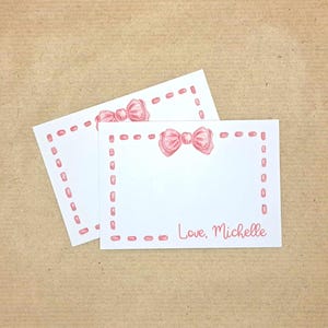 May include: Two rectangular white note cards featuring a pink hand-drawn bow at the top centre and a pink dashed border. The front card displays the text Love, Michelle in a pink script font, while the second card is partially visible behind it.