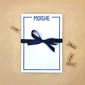 May include: A rectangular white notepad featuring a dark blue hand-drawn border and the name MOISHE printed in dark blue at the top. The notepad is tied horizontally with a dark blue satin ribbon tied into a bow at the center of the page.