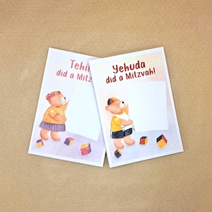 May include: Two rectangular greeting cards featuring watercolor illustrations of teddy bears. One card shows a bear with the text 'Tehila did a Mitzvah!' and the other shows a bear with the text 'Yehuda did a Mitzvah!', both with colorful blocks.