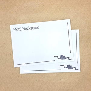 May include: Two rectangular white cards featuring the text Motti Heckscher in the top left corner. Each card has a thin black border line along the right side and bottom, with a small graphic of two stacked boxes and a ribbon in the bottom right corner.
