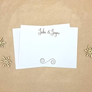May include: Two white rectangular note cards featuring the black printed text John & Jayne at the top and a decorative black swirl flourish at the bottom. Three small, light brown wooden flower-shaped embellishments are placed around the cards.