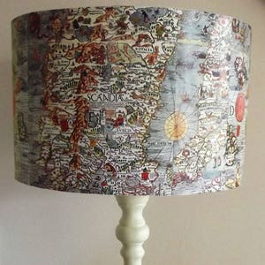 May include: A white table lamp with a cylindrical lampshade featuring an antique map of Scandinavia. The map is printed in black, brown, red, and blue ink. The lamp base is a simple, white, turned wood design.