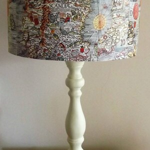 May include: A white table lamp with a cylindrical lampshade featuring an antique map of Scandinavia. The map is printed in black, brown, red, and blue ink. The lamp base is a simple, white, turned wood design.