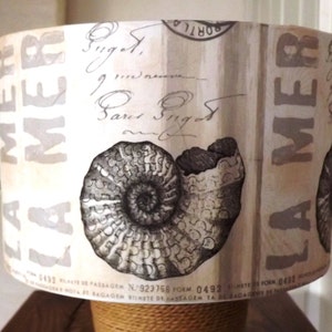 May include: A white lampshade featuring a black and white nautilus shell design. The lampshade has the text "LA MER" printed on it in a grey font.