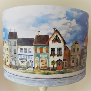 May include: A lampshade featuring a watercolour design of a row of colourful houses against a blue sky with white clouds. The houses have windows, doors, and signs that read "Bakery" and "Flowers".