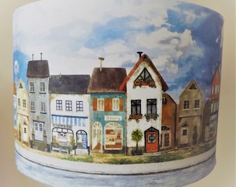 May include: A lampshade with a watercolor design of a row of colorful houses with blue sky and white clouds. The houses have windows, doors, and signs that say "Bakery" and "Flowers".