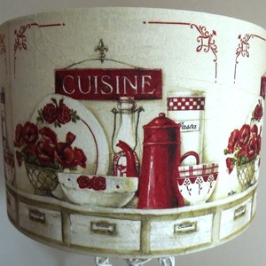 May include: A cream-coloured lampshade with a red and white kitchen-themed design. The design features a sign that says "Cuisine", red flowers, and various kitchen utensils. The lampshade has a row of drawers with labels that say "Pasta", "Spices", and "Cuisine".