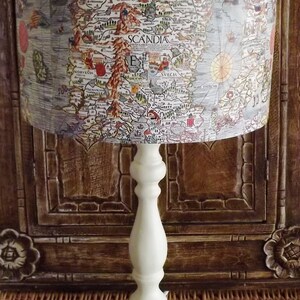 May include: A white table lamp with a cream-colored shade featuring an antique map of Scandinavia. The map shows the countries of Norway, Sweden, and Finland. The lamp is sitting on a woven wicker surface.