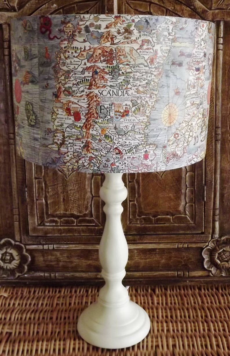 May include: A white table lamp with a cream-colored shade featuring an antique map of Scandinavia. The map shows the countries of Norway, Sweden, and Finland. The lamp is sitting on a woven wicker surface.