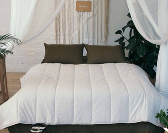 May include: A white duvet cover with a quilted design, on a bed with two olive green pillows. The bed is in a bedroom with white walls and a white sheer canopy. There are plants in the room and a wooden crib in the background.