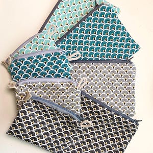 Puede incluir: Una colección de estuches con cremallera de varios tamaños, con un patrón repetido en forma de abanico. Los estuches muestran una gama de colores, incluyendo verde azulado, gris, negro y dorado. Cada estuche tiene una cremallera en contraste y un pequeño lazo de tela.