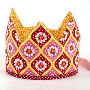 Op de afbeelding: Een roze, gele en rode stoffen kroon met een wit madeliefjespatroon. De kroon heeft een witte stippenrand.