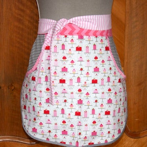 Op de afbeelding: Een roze en witte schort met een geruit patroon en een cupcake-ontwerp. De schort heeft een roze en witte gestreepte tailleband en een strik.