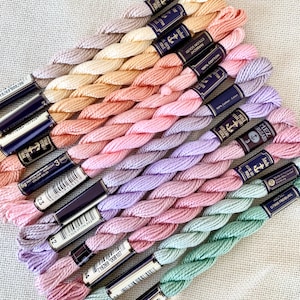 Könnte beinhalten: Eine Auswahl an Anchor-Stickgarn in Pastelltönen wie Rosa, Lavendel, Pfirsich und Mintgrün. Jede Strähne ist mit dem Markennamen und Farbinformationen versehen. Das Garn besteht aus 100 % Baumwolle.