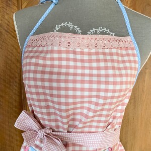Può includere: Un grembiule a quadri rosa e bianco con un nastro a pois blu e bianchi. Il grembiule ha un bordo in pizzo e una tasca con una stampa floreale blu e bianca con cupcake e ciliegie.