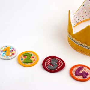 Può includere: Cinque cerchi di feltro con i numeri da 1 a 4. I cerchi sono in linea su una superficie bianca. Una corona di feltro gialla con pois bianchi si trova nell'angolo in alto a destra dell'immagine.