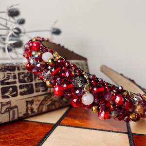 May include: A headband densely embellished with a variety of red, gold, and clear beads. The accessory features a mix of faceted, round, and smooth beads in varying sizes, creating a textured surface across the entire band structure.