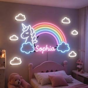 May include: A neon sign in a child's bedroom, featuring a unicorn, rainbow, and clouds, with the name "Sophia" in pink. The sign is mounted on a grey wall above a pink bed. Additional cloud-shaped lights surround the main sign, creating a whimsical theme.