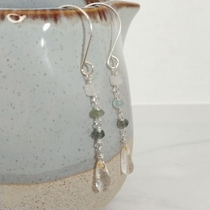 May include: A pair of silver dangle earrings with a teardrop-shaped clear quartz crystal at the bottom. The earrings feature a chain with small green and blue gemstones.