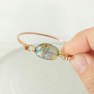May include: A copper wire bangle bracelet with a single oval-shaped labradorite gemstone. The gemstone has a iridescent blue and green sheen.