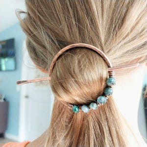 May include: Copper hair pin with a half-circle design and a straight pin. The hair accessory features a strand of teal-colored beads wrapped in copper wire. The hair pin is inserted into a bun of blonde hair.